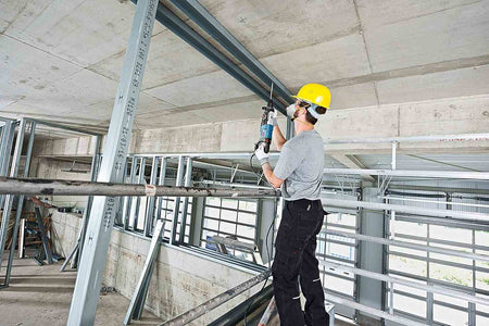 An electrician using the Bosch Bulldog rotary hammer to drill into a steel-reinforced concrete ceiling structure from a scaffold, demonstrating its power and reach.