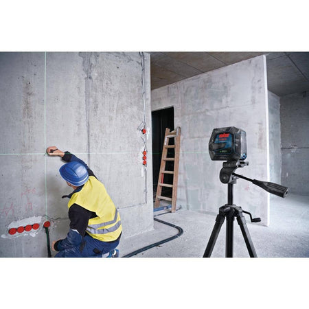 An application photo of the Bosch GCL100-40G mounted on a tripod in a construction setting, where a professional contractor is using the projected green laser lines to accurately mark layout points on a concrete wall.