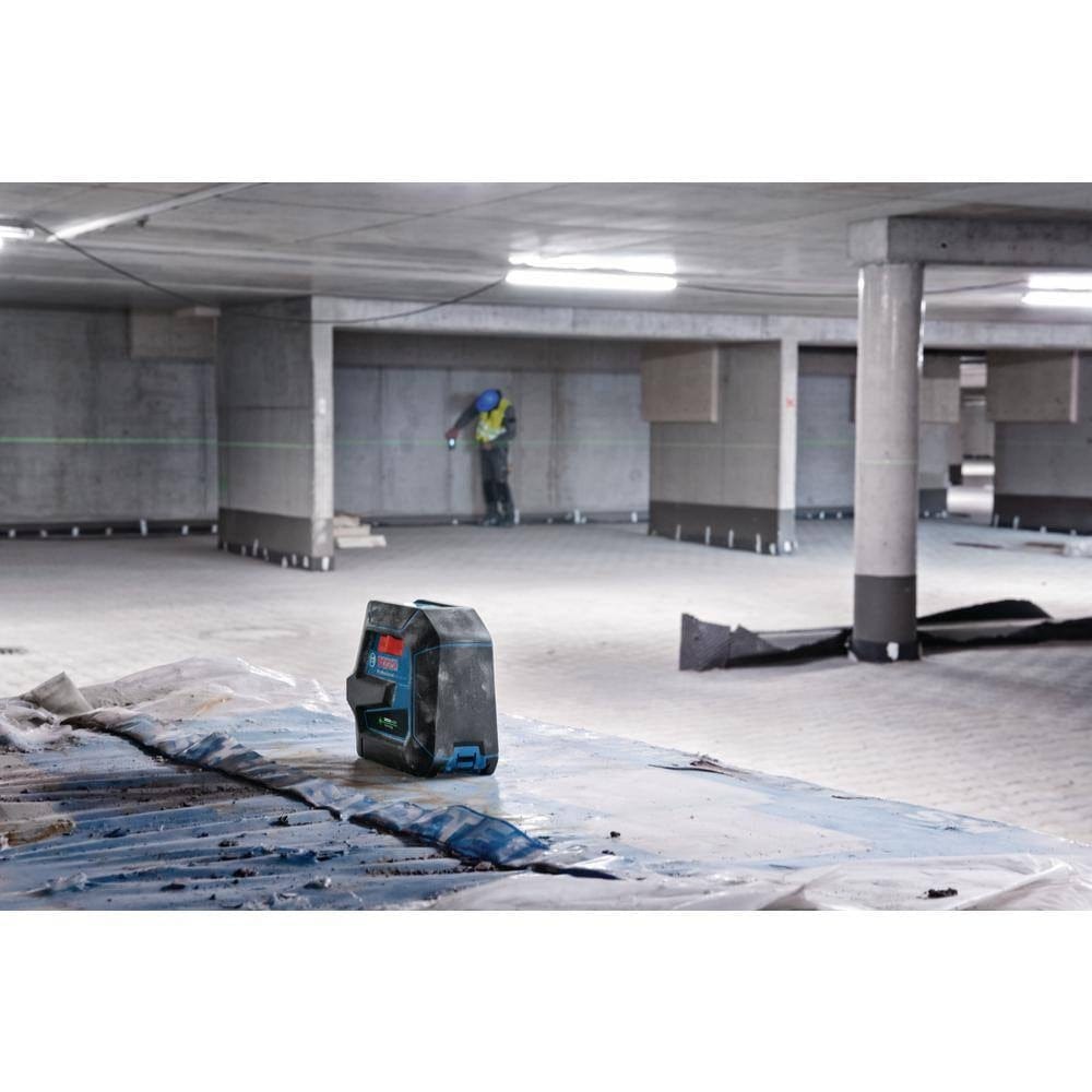 An indoor jobsite action shot showing the Bosch green beam laser level sitting on a flat surface, projecting a long-distance horizontal leveling line across a large open basement for wall and electrical installation.