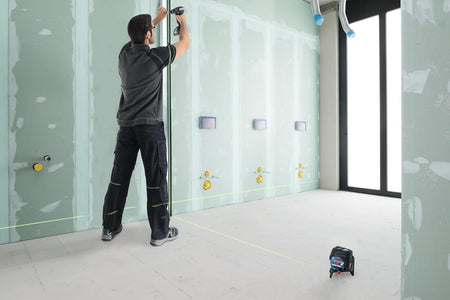 A professional fastening a metal stud track to a wall, using the vertical green beam projected from the floor-mounted laser to ensure the installation is perfectly plumb.