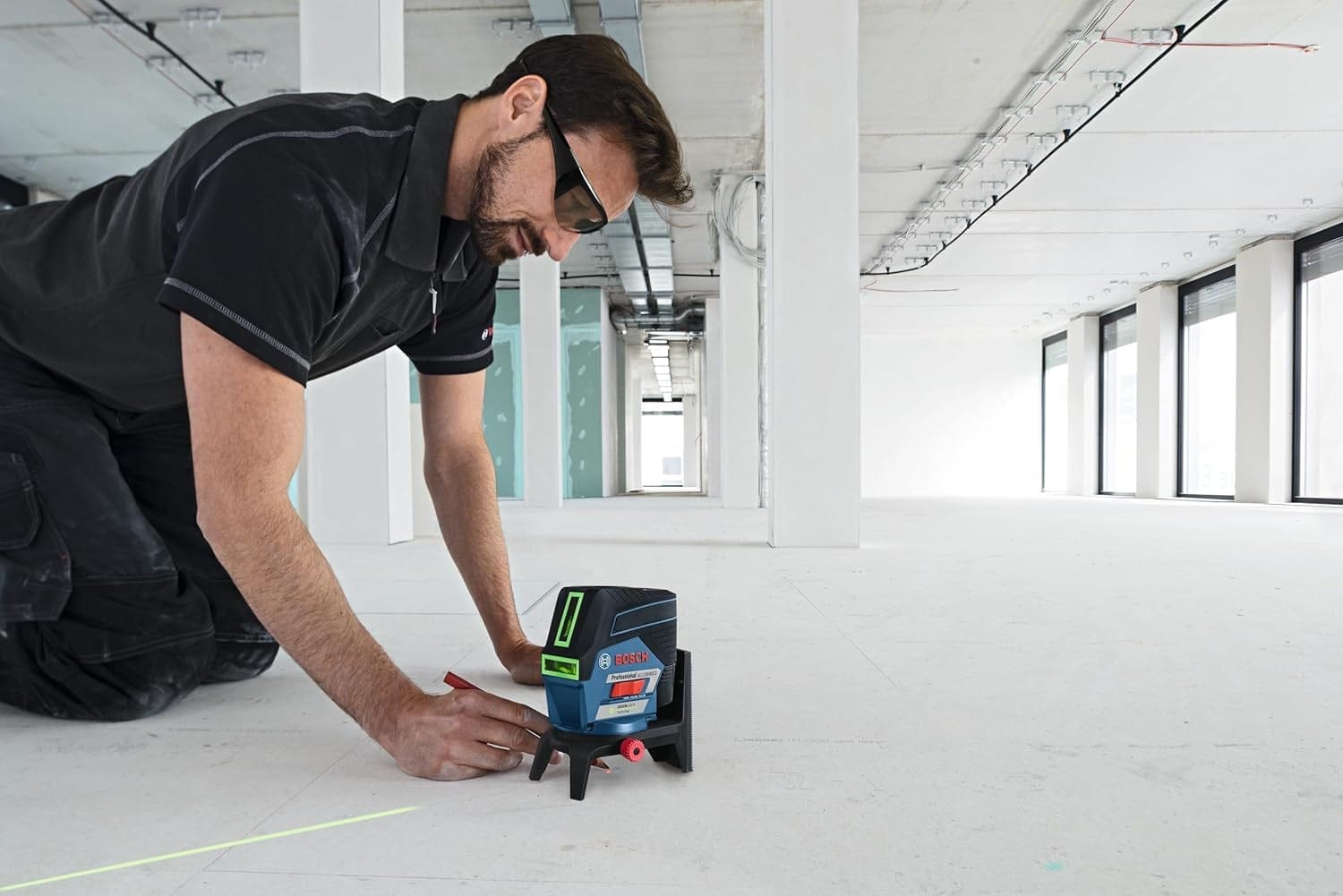 A close-up of a worker kneeling on the floor and using a pencil to mark a reference point directly underneath the laser unit, utilizing the tool's plumb down feature.