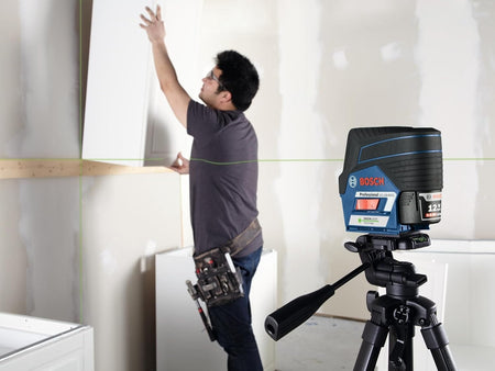 A carpenter installing white cabinetry using the laser level mounted on a tripod to project a precise horizontal leveling line.