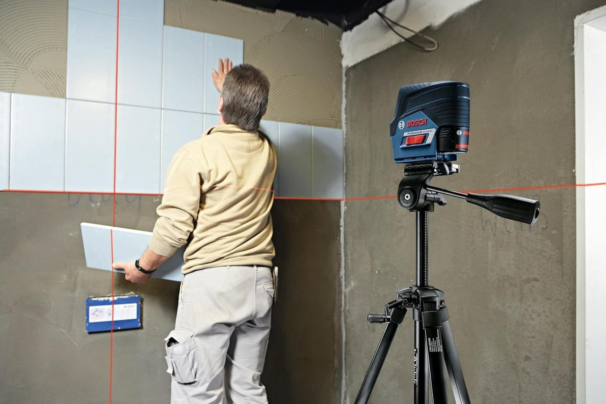 Professional tile installer using the Bosch GCL100-80C cross-line laser level on a tripod to ensure perfect vertical and horizontal grout line alignment on a wall.