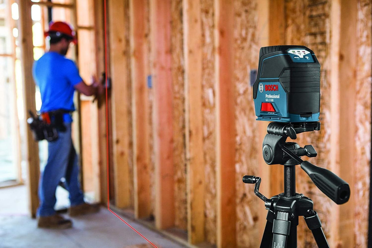 Bosch GCL 2-160 laser level mounted on tripod projecting red vertical line onto wooden framing; worker aligns measurements in background.