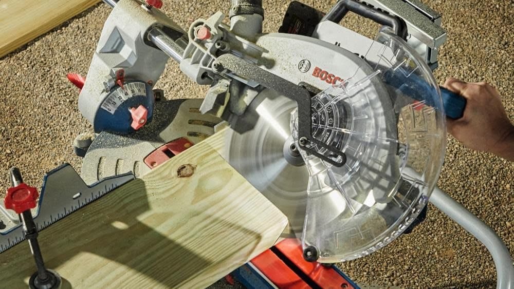 An overhead action shot of the Bosch Surgeon miter saw performing a sliding crosscut on a large wooden plank at an outdoor jobsite, emphasizing its cordless mobility.