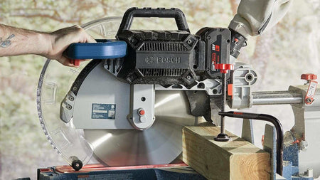 An action shot of the Bosch GCM18V-12SDN miter saw cutting through a thick 4x4 structural wood post, demonstrating the high-torque BITURBO brushless motor and material clamp in use.