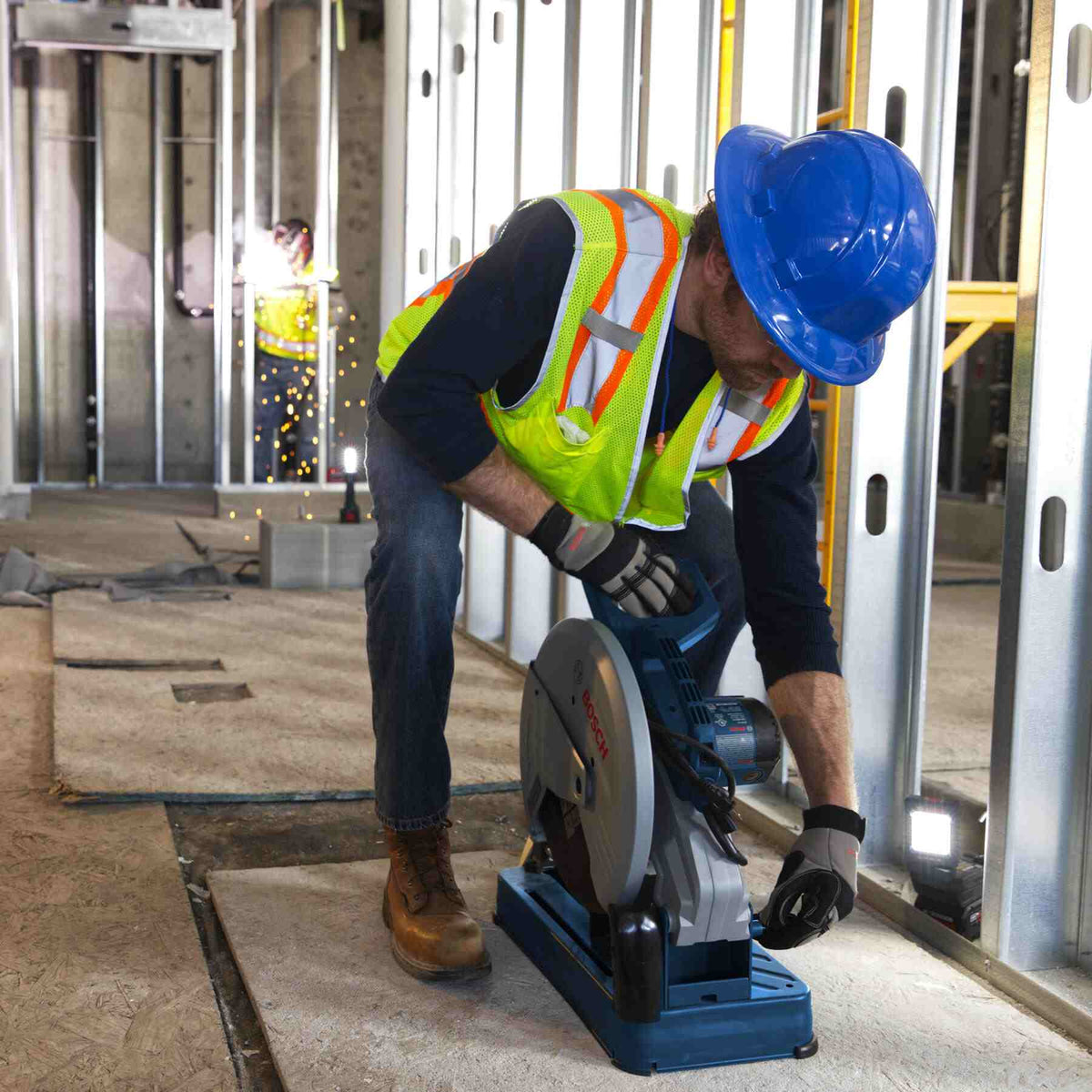 A professional wearing a blue hard hat and safety vest using the chop saw to cut metal framing studs in a commercial construction environment.