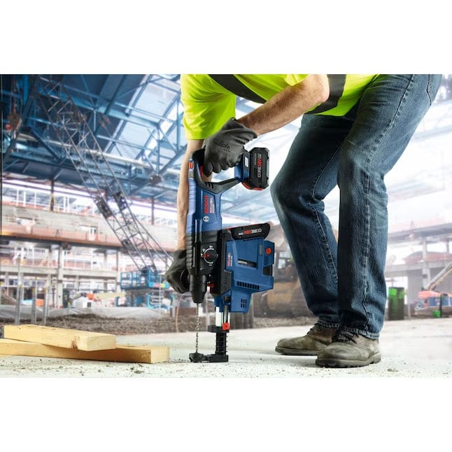 Worker in high-visibility gear using a Bosch rotary hammer vertically on a concrete surface at a large construction site.