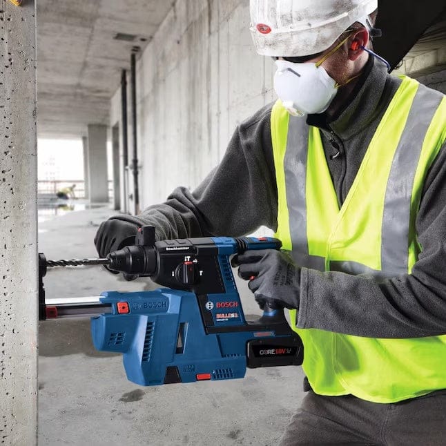 Construction worker using a Bosch GBH 18V-45 C rotary hammer drill on a concrete wall while wearing full safety gear.