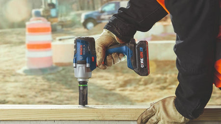 Close-up action shot of a worker using the 18V mid-torque impact wrench and a green-banded impact socket to drive a heavy-duty fastener into a wooden structural beam outdoors.