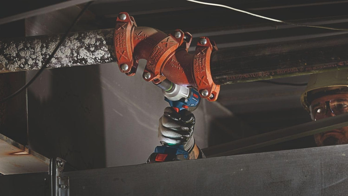 An action shot showing a worker using the Bosch impact wrench to tighten hardware on a large overhead industrial pipe assembly.