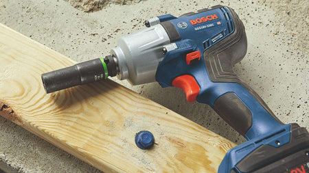 A high-angle product shot of the Bosch mid-torque impact wrench resting on a wooden board next to a blue heavy-duty bolt, showing an impact socket attached to the anvil.