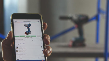 A close-up shot of a hand holding a smartphone displaying the Bosch Toolbox App, which is connected to the GDS18V-330C impact wrench to customize modes and settings like RPM adjustment.