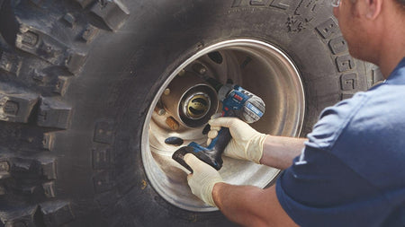 Close-up action shot of the cordless mid-torque impact wrench being used by a worker in safety gloves to remove or tighten a lug nut on a large vehicle wheel.