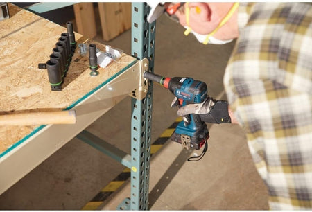 Action shot showing a professional using the Bosch impact tool with a deep socket to secure a metal bracket to an industrial storage rack, with a set of impact sockets visible on a nearby workbench.
