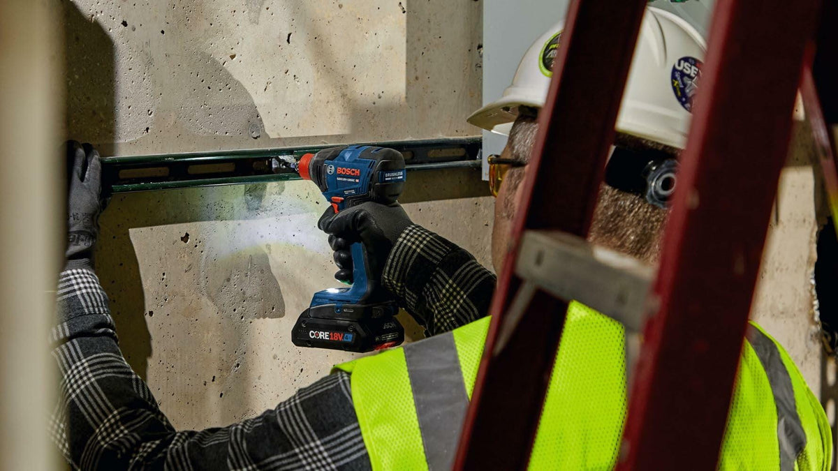 A worker using the Bosch GDX18V-1860CN impact driver (with a drill bit) to fasten into a concrete wall, holding a level in the opposite hand to ensure accuracy.