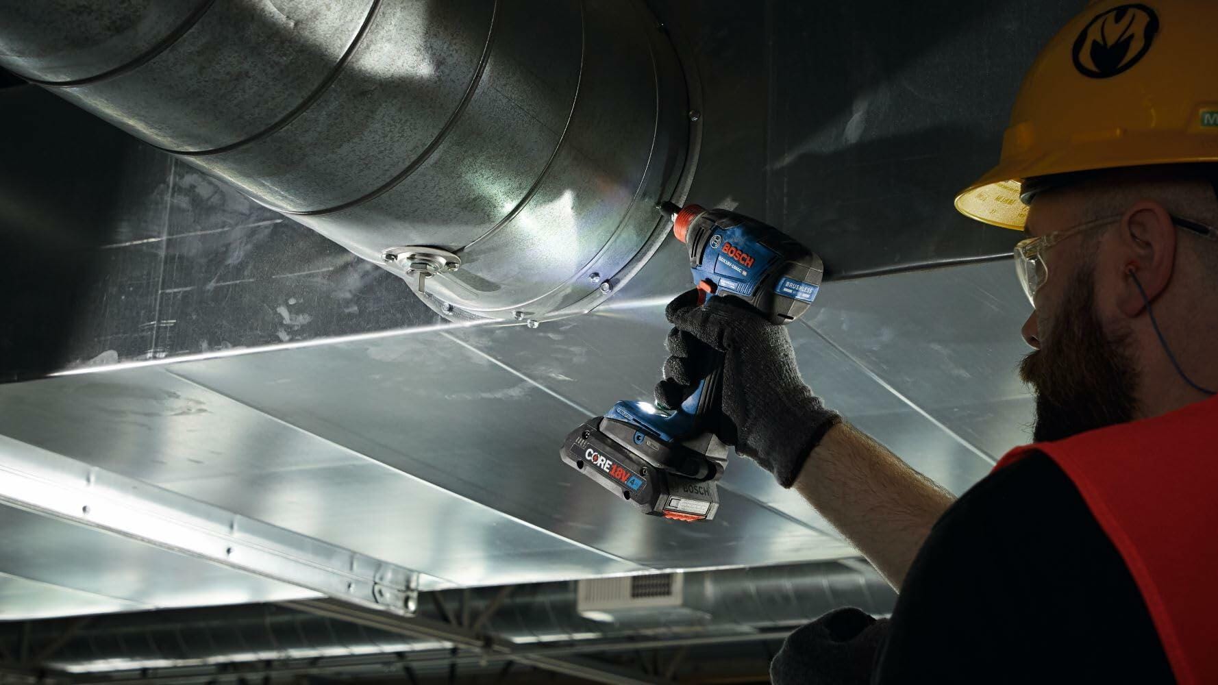 A worker wearing safety gear uses the Bosch GDX18V-1860CN impact driver (with an attached battery) to fasten components into a large piece of ceiling HVAC ductwork.