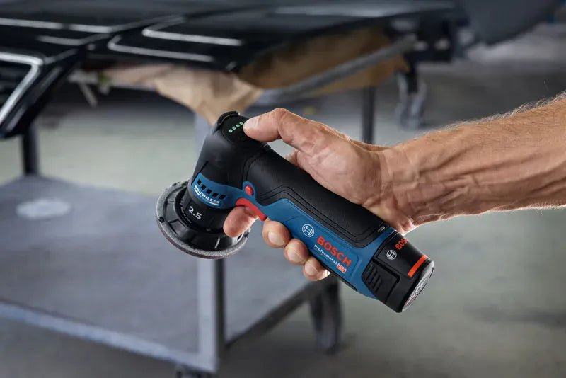 A close-up view of a technician's hand holding the Bosch GEX12V-77N 12V Max random orbital sander from the side, with the thumb resting on the integrated top-mounted 5-speed digital user interface. The battery status indicator lights glow green, showing immediate operational readiness for professional car paint repair.