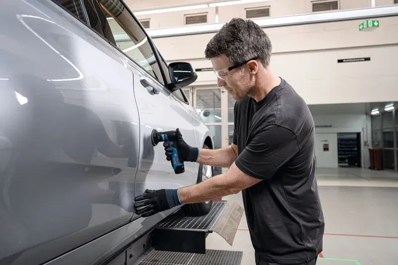 An automotive paint reconditioning specialist wearing clear safety glasses and black protective gloves operating the compact Bosch 12V random orbital sander bare tool. Pictured inside a clean commercial factory floor or bodyshop, the technician holds the sander vertically with one hand to sand and blend clear coat paint imperfections on a silver car door panel.