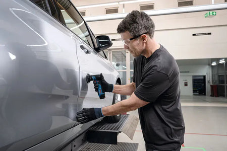 An automotive paint reconditioning specialist wearing clear safety glasses and black protective gloves operating the compact Bosch 12V random orbital sander bare tool. Pictured inside a clean commercial factory floor or bodyshop, the technician holds the sander vertically with one hand to sand and blend clear coat paint imperfections on a silver car door panel.