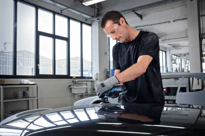 A comprehensive workshop view of a refinishing specialist bent over a gloss black car surface, carefully operating the compact Bosch GEX12V-77N handheld brushless sander. The backdrop highlights a bright, professional auto detailing garage with panoramic windows illuminating the workpiece.