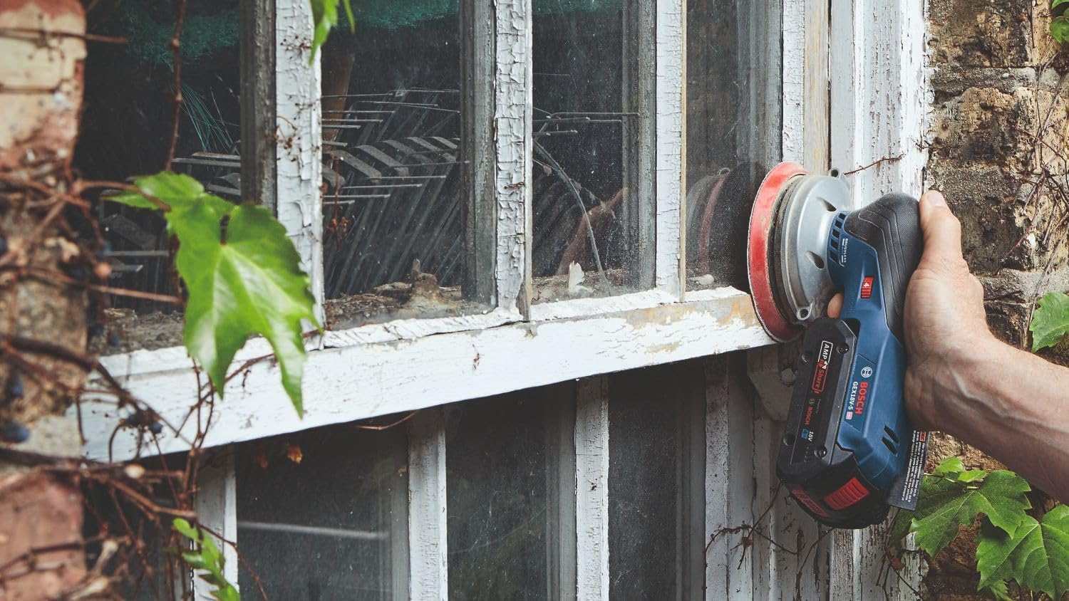 A person using the Bosch 18V brushless 5-inch random orbit sander to perform vertical sanding on a weathered window frame, showcasing the tool's compact shape and control.