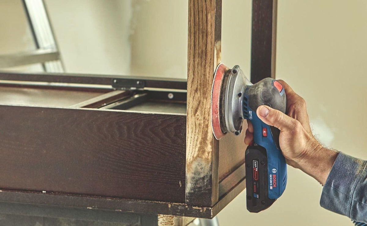 A woodworker using the Bosch cordless random orbit sander to refine the finish on the vertical leg of a dark wood furniture piece in a workshop.