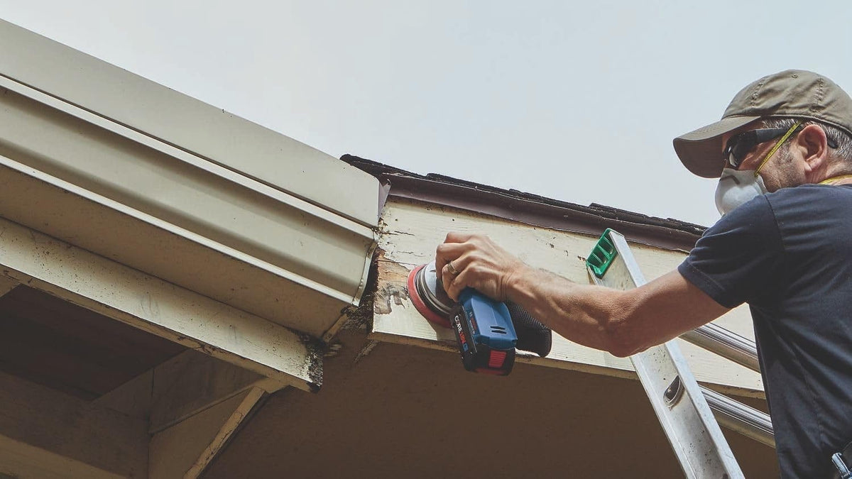 A contractor on a ladder using the Bosch 18V random orbit sander to perform repair sanding on a wooden roof soffit and fascia board near a gutter system.