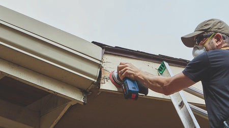 A contractor on a ladder using the Bosch 18V random orbit sander to perform repair sanding on a wooden roof soffit and fascia board near a gutter system.