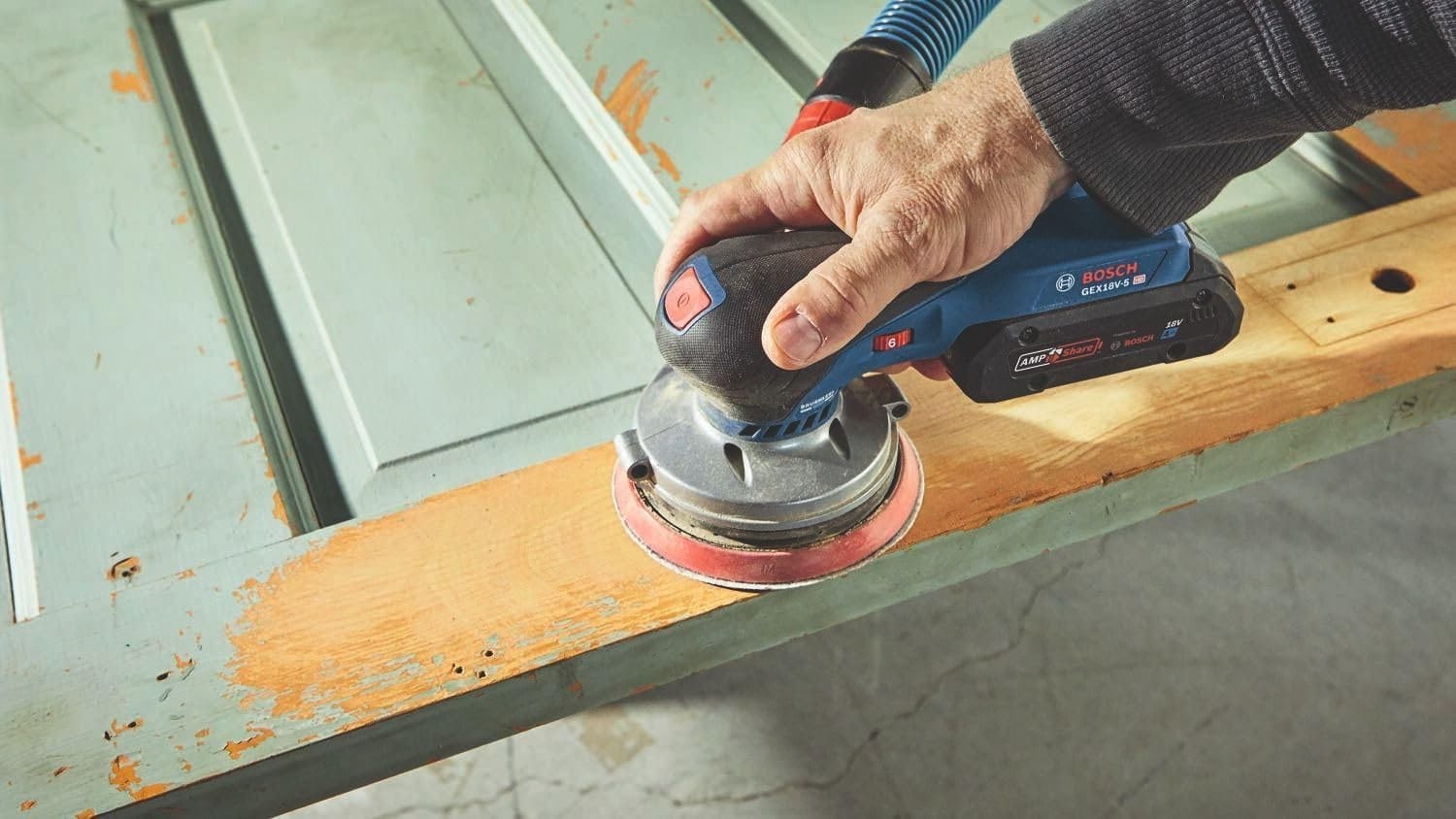 A person using the Bosch brushless sander with a vacuum attachment to strip old blue paint from a flat wooden door panel, revealing the natural wood grain.