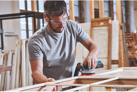 A professional utilizing the Bosch 18V brushless random orbit sander with an attached vacuum hose to perform smooth, dustless finish sanding on a large wooden door frame.