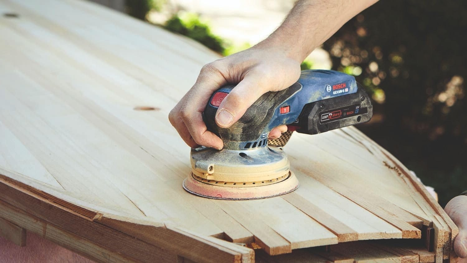 A close-up action shot of a worker using the Bosch cordless palm sander to refine the surface of a wooden surfboard, demonstrating the tool's precision on contoured workpieces.