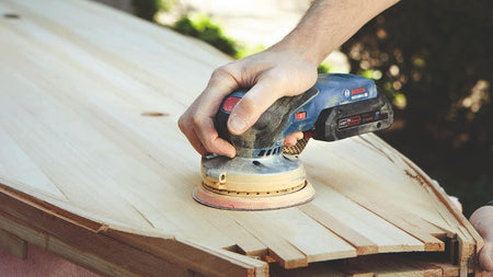 A close-up action shot of a worker using the Bosch cordless palm sander to refine the surface of a wooden surfboard, demonstrating the tool's precision on contoured workpieces.