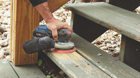 An on-site view of a contractor using the Bosch 18V random orbit sander with a dust bag to strip and smooth old paint from exterior wooden stairs.