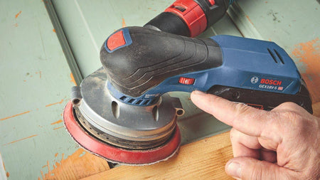 A detailed view of a user's hand adjusting the red variable speed selector on the Bosch GEX18V-5 sander to match the OPM to the specific wood material.