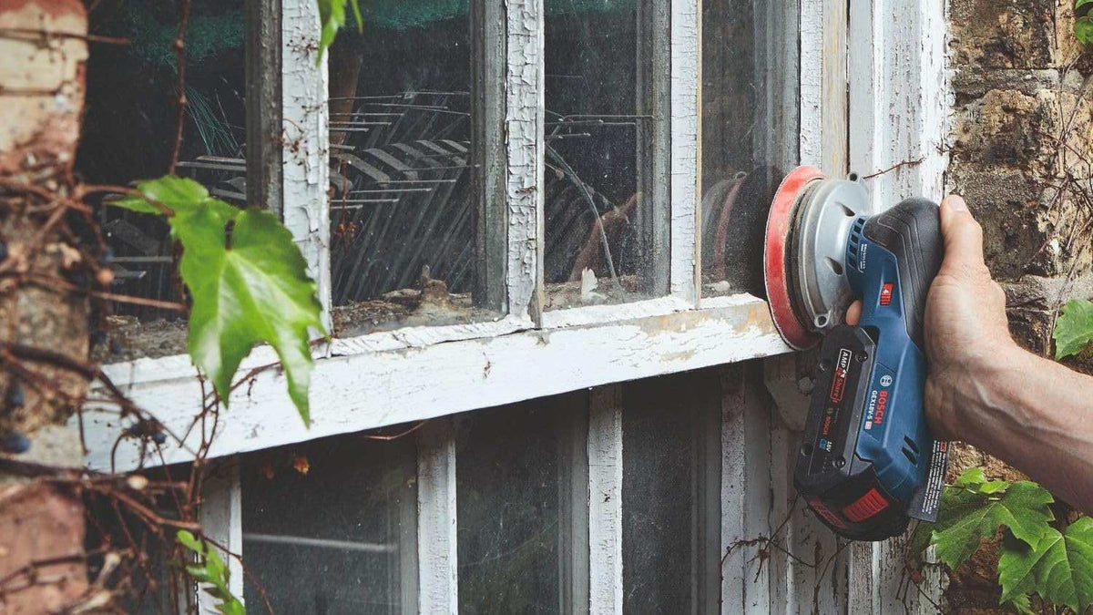 A user holding the sander vertically to remove paint from an exterior wooden window frame, demonstrating the tool's compact control.