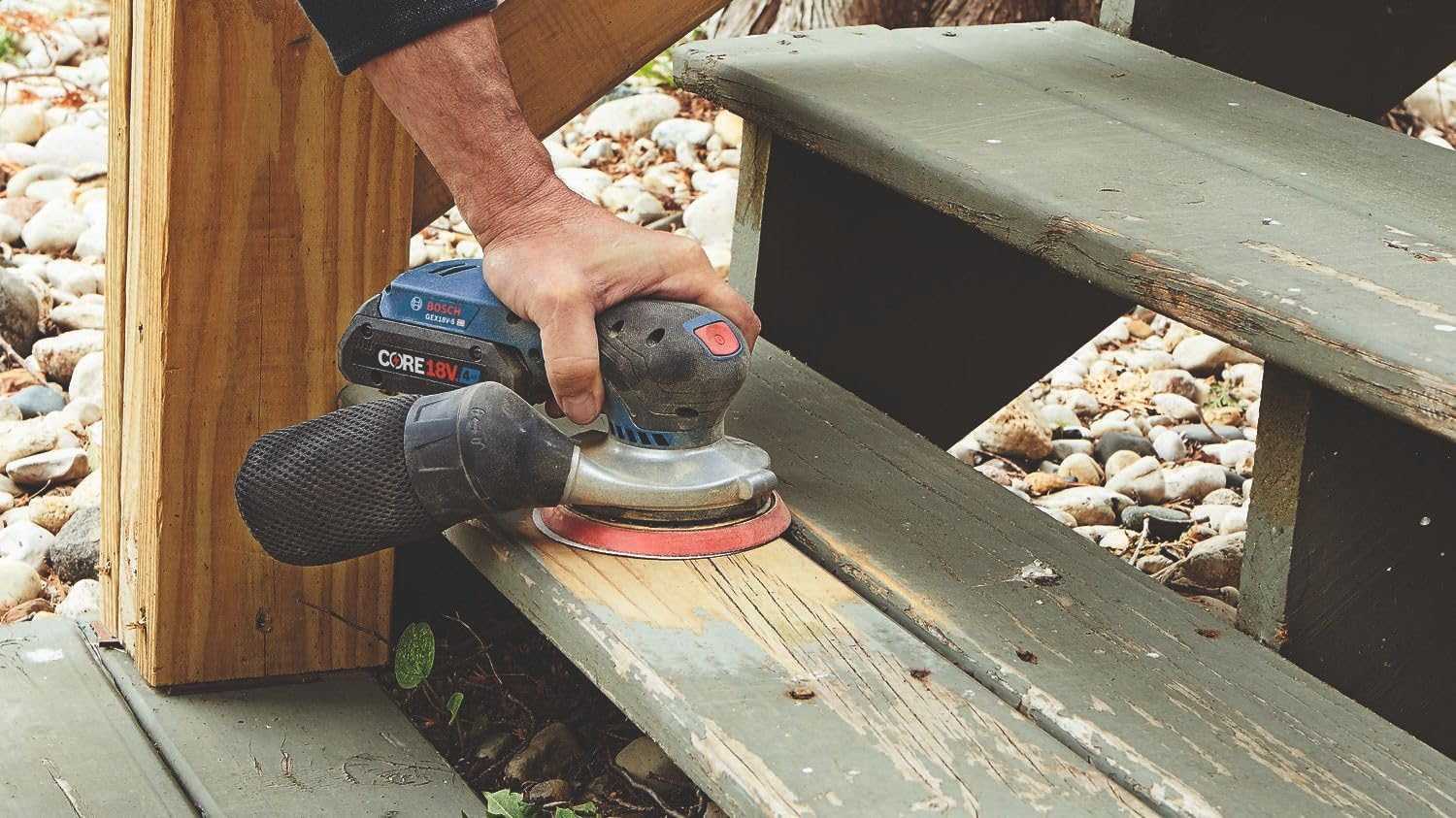 Close-up of a user's hand guiding the cordless sander with the dust bag attached while smoothing a wooden step surface.