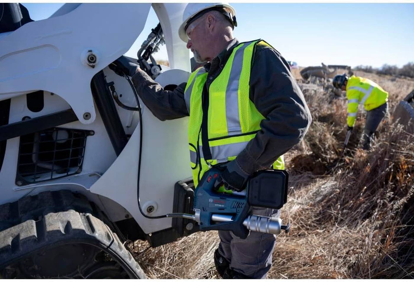 An operator wearing a safety vest and hard hat uses the Bosch GFP18V-10N with the included shoulder strap to lubricate the pivot points of a white compact track loader on a job site.