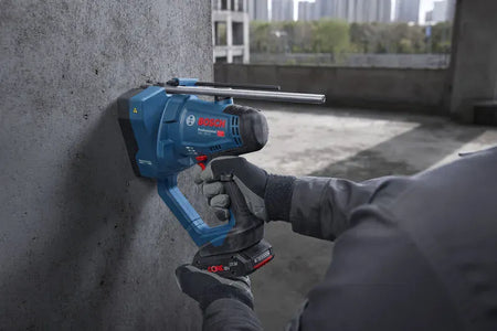 An active horizontal field shot capturing a trade professional operating the Bosch GGC18V-12N 18V max brushless rod cutter to punch an anchor hole guideline into a solid concrete masonry wall. The uncropped action shot details the surrounding commercial building framework and highlights the stable two-hand control layout of this essential tool.