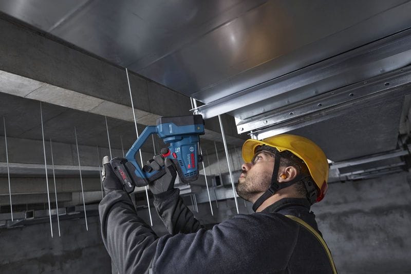 A low-angle field action shot showing an electrical or mechanical installer looking upward while utilizing the Bosch GGC18V-12N 18V max rod cutter to trim overhead support drops. Pictured inside a raw concrete commercial building ceiling space, the worker easily maneuvers the tool to cleanly slice through hanging zinc-plated fasteners without generating dangerous jobsite sparks.