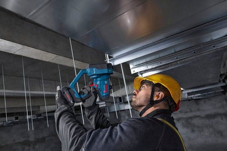 A low-angle field action shot showing an electrical or mechanical installer looking upward while utilizing the Bosch GGC18V-12N 18V max rod cutter to trim overhead support drops. Pictured inside a raw concrete commercial building ceiling space, the worker easily maneuvers the tool to cleanly slice through hanging zinc-plated fasteners without generating dangerous jobsite sparks.