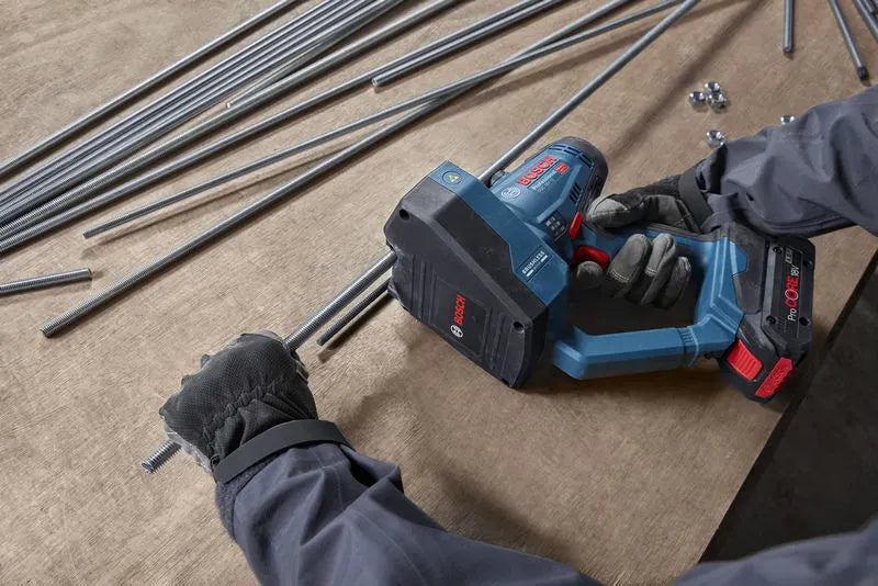 An uncropped high-angle view identical to file GGC18V-12N 5.jpg showing a craftsman's gloved hands aligning a mild steel fastener rod across the indexing jaw assembly of the Bosch GGC18V-12N cordless shear tool. A bundle of loose steel threaded rod lengths rests on the wooden shop workbench background, emphasizing the tool's high-volume pre-fabrication capabilities.