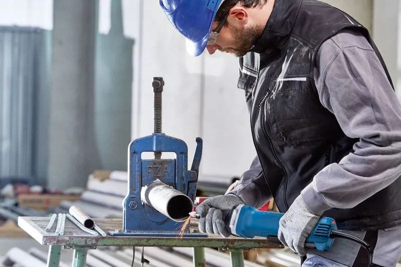A front-facing action shot of a workshop technician wearing a blue hard hat and safety glasses guiding the extended nozzle of the Bosch paddle-switch die grinder to polish the interior edge of a metal pipe clamped into a structural pipe vice.