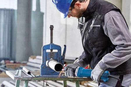 A front-facing action shot of a workshop technician wearing a blue hard hat and safety glasses guiding the extended nozzle of the Bosch paddle-switch die grinder to polish the interior edge of a metal pipe clamped into a structural pipe vice.