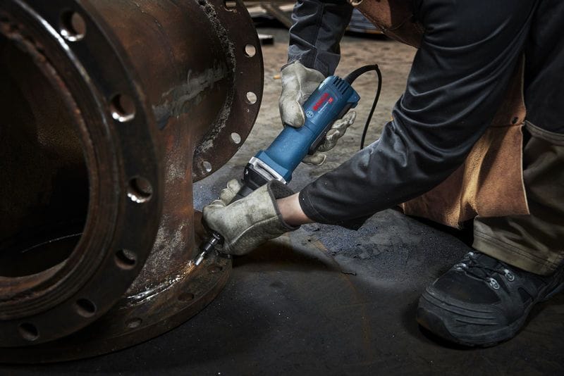 An industrial application shot showing a technician in leather work gloves using the long-nose Bosch straight grinder to polish the machined internal surface of a heavy steel pipe joint section, with an attached grinding point bit visible inside the collet chuck.