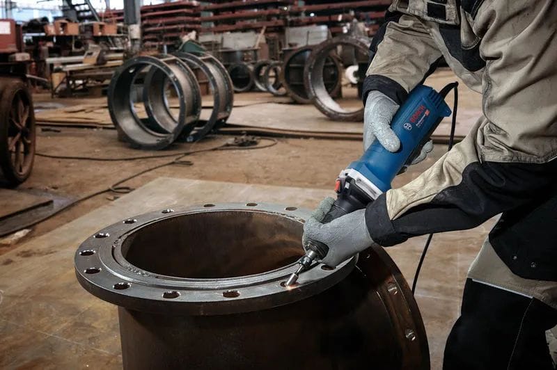 A wide workshop perspective shot of an industrial metalworker wearing heavy safety gloves using the Bosch corded straight die grinder to mill and smooth the inner rim of a large circular steel fabrication flange.
