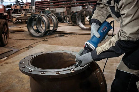 A wide workshop perspective shot of an industrial metalworker wearing heavy safety gloves using the Bosch corded straight die grinder to mill and smooth the inner rim of a large circular steel fabrication flange.
