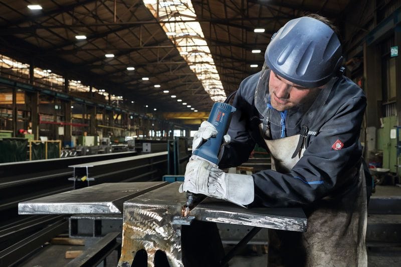 A high-angle industrial action shot of a professional welder wearing a protective safety face shield helmet and thick leather apron using the Bosch long-nose die grinder to bevel a thick steel beam plate seam, generating a concentrated cluster of orange sparks.