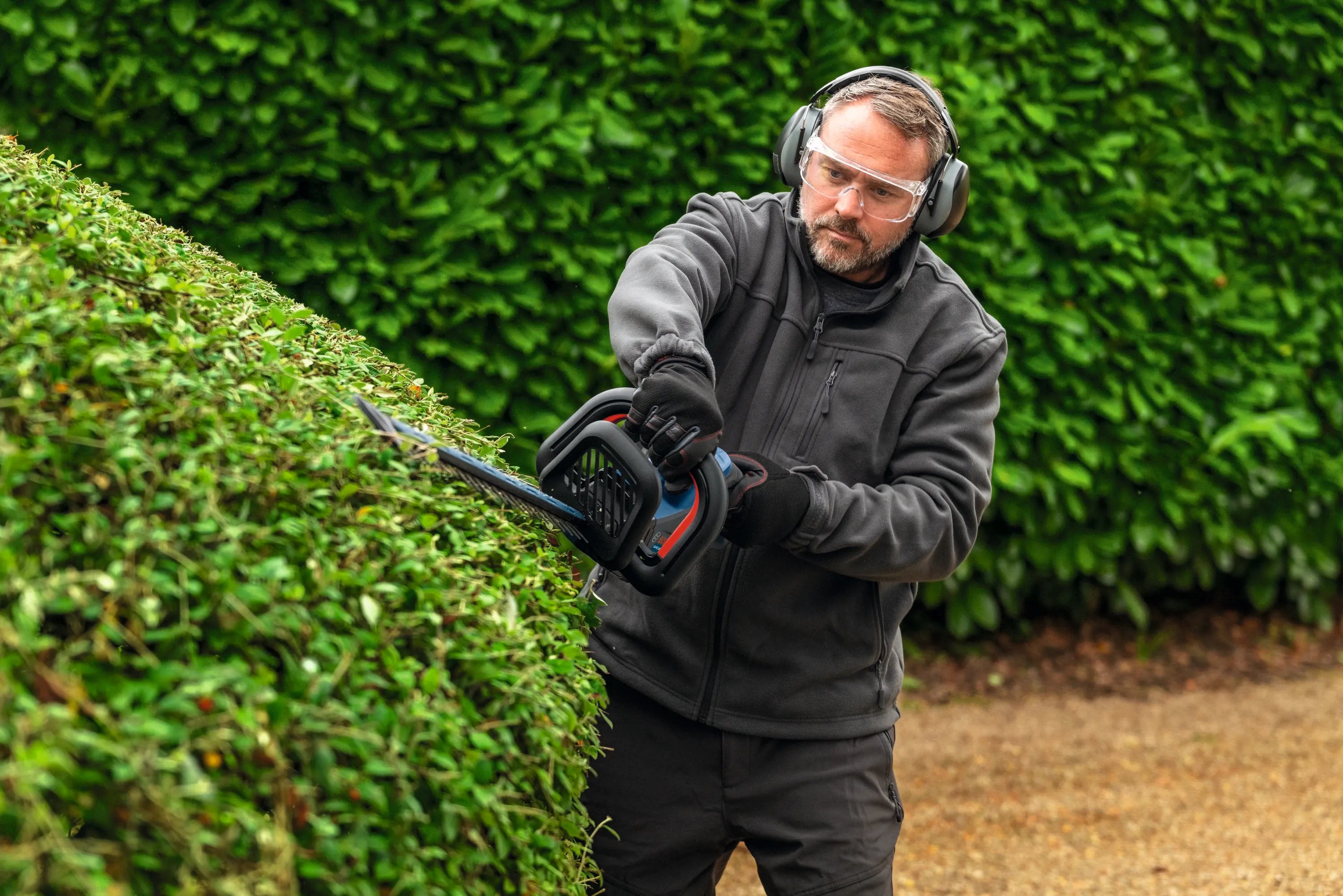 A front-facing action shot of a professional landscaper wearing eye protection and noise-dampening earmuffs as he arches a cordless Bosch trimmer smoothly over a large rounded hedge dome to sculpt a precise, clean landscape border.