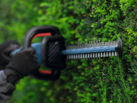 A close-up action shot focusing on the silver reciprocating steel teeth of a 24-inch dual-action hedge trimmer blade as a worker wearing protective gloves guides it horizontally through dense green shrub leaves.